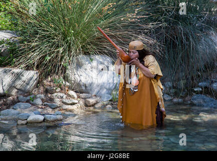 Traditional California Native American woman of the Acjachemen Nation (San Juan Capistrano Mission Indians) dressed in deer hides, shell beads and basket hat drinks from a spring using a large gourd dipper. (MR) Stock Photo