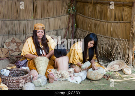 Acjachemen Nation Family (Mission Indians) grind acorns inside a furnished Tule reed hut, traditional housing of the Indians of Southern California, located in the Tongva village, Santa Fe Springs CA (MR) Stock Photo