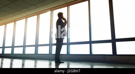 Full length shot of businessman standing near office window and talking on mobile phone. Man looking outside the window and making a phone call. Stock Photo