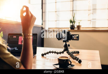 Female vlogger looking at camera while editing video on her laptop. Young woman working on computer with coffee and camera on her desk. Stock Photo