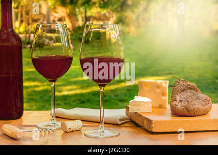 Two glasses of red wine and a bottle, with slices of cheese at a picnic, on a wooden table, with green trees and grass in the blurred background, with Stock Photo