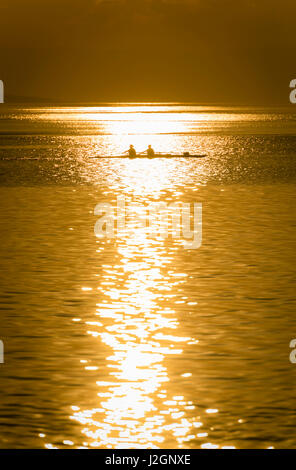 A team of two oarswomen is training in their rowboat on Lake Geneva, Switzerland. Back-lit, evening sunset. Stock Photo