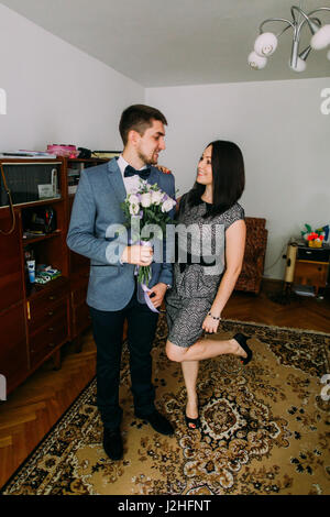 Handsome man holding flowers posing indoors with pretty young woman, playfully raises her leg. Stock Photo
