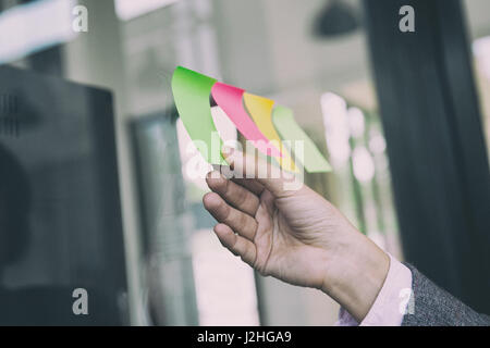 Female business person hand holding blank sticky paper note on glass wall in office Stock Photo