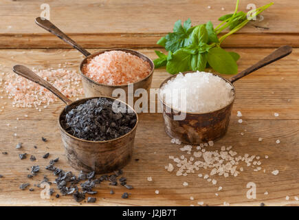 Three different types of salt in silver containers on a worn out wooden surface. White sea salt, pink Himalayan and black Maldon. Stock Photo