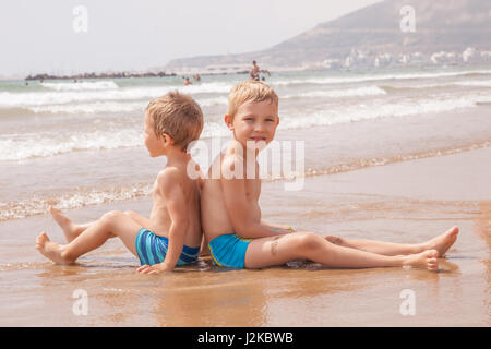 Two cute adorable funny little brothers boys sitting on the beach ocean sea travel