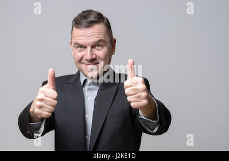 Grinning man in dark suit jacket with two thumbs up over gray background. Includes copy space. Stock Photo