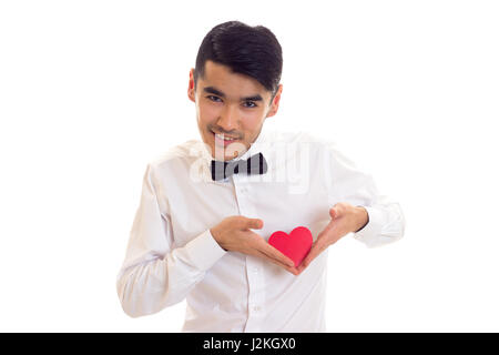 Smiling man with black hair in white T-shirt with black bow-tie holding a red paper heart on white background in studio Stock Photo