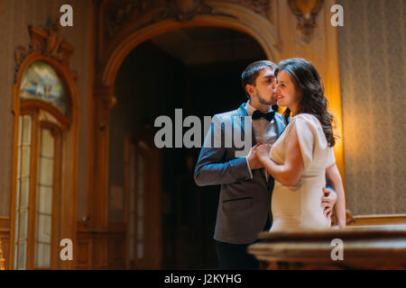 Beautiful wedding couple embracing and kissing near balustrade at gorgeous antique stairs with the background of royal wooden vintage interior Stock Photo