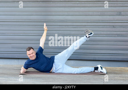 Man stretching himself on a mat, laying on his side with one leg and hand raised, looking at camera and smiling Stock Photo