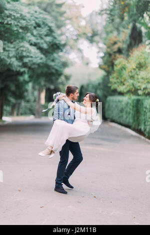 Beautiful wedding couple portrait with groom carrying his smiling bride on green park lane Stock Photo