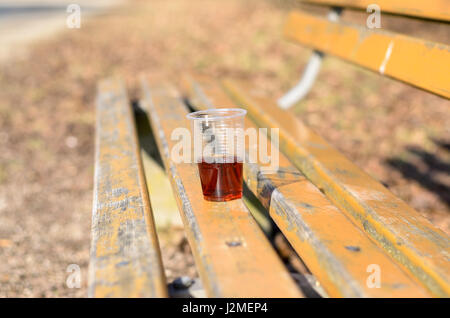 Close-up shot of plastic cup with red vine or other beverage on old painted yellow park bench in autumn sunny day Stock Photo