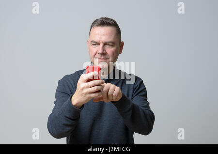 Man in his middle age wearing black sweater looking at red cellphone in his hands, standing against plain grey background Stock Photo
