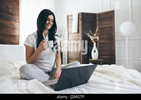 Relaxing on the bed. Beautiful young woman enjoying a cup of coffee and using her laptop Stock Photo