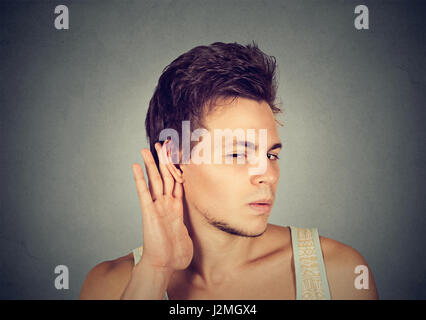 Closeup portrait a man placing hand on ear listening carefully to gossip isolated on gray wall background. Human emotion Stock Photo