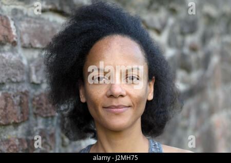 Single pretty young adult black woman with long hair smiling near gray brick wall Stock Photo