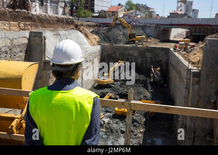 Road construction engineer monitors excavators (baggers) working digging for a subway. Dressed in an working uniform and protective white helmet. From Stock Photo