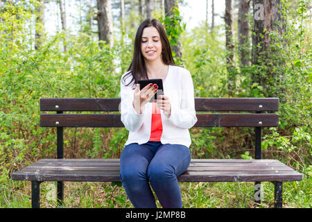 Attractive young woman dressed casually uses e-book/tablet sitting on a wooden bench in the forest. Modern lifestyle using portable mobile devices eve Stock Photo