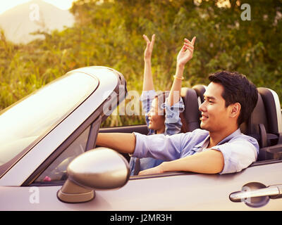 young asian couple riding in a convertible sport car enjoying the cool breeze at sunset. Stock Photo