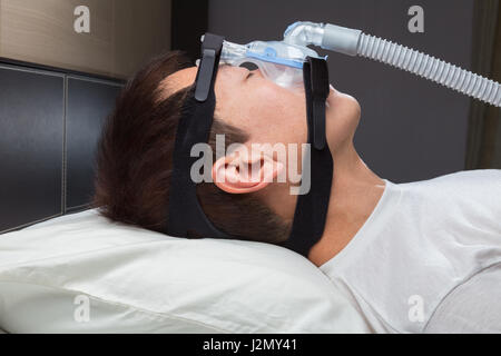 Asian man with sleep apnea using CPAP machine, wearing headgear mask connecting to air tube Stock Photo