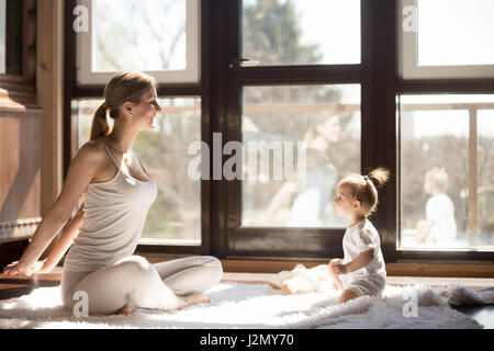 Young yogi mother at Easy Seat pose, cute baby daughter sitting near her, starting a new sporty day together, enjoying morning exercises at home, fami Stock Photo
