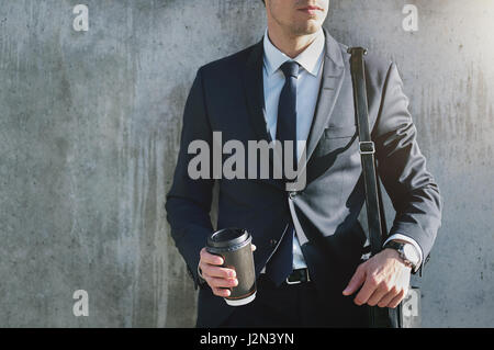 A businessman wearing stylish suit with the coffee cup standing near the gray wall. Stock Photo