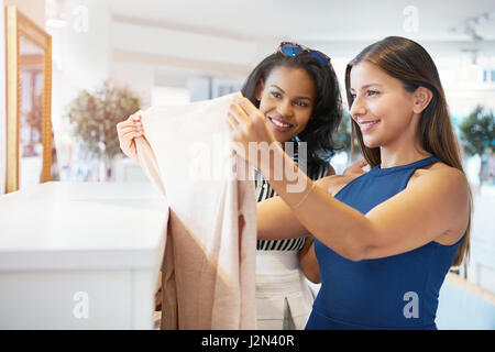 Two young female friends choosing clothes together in a fashion boutique discussing a garment holding it up to the light for inspection Stock Photo
