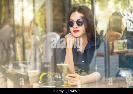 young asian woman holding her smartphone in a coffee shop seen through glass window with reflection on a bright sunny day Stock Photo