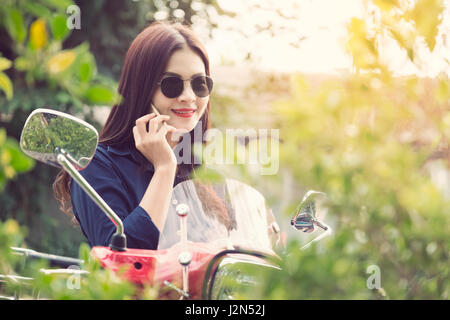 Young happy asian woman talking on her phone at her red motorcycle, vintage retro color style with sun flare Stock Photo