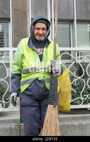 Tehran Province- IRAN-March 17, 2016 A Portrait of An Old Urban Street Cleaner Man Holding Hand-made Sweeping Tool By Gloved Hand in Front of Modern B Stock Photo
