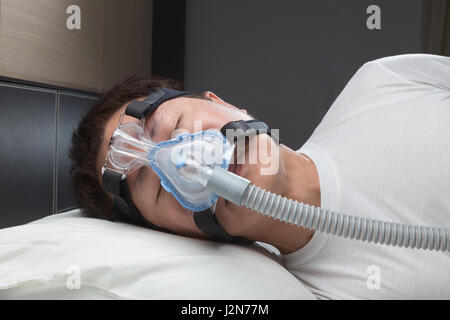 Asian man with sleep apnea using CPAP machine, wearing headgear mask connecting to air tube Stock Photo