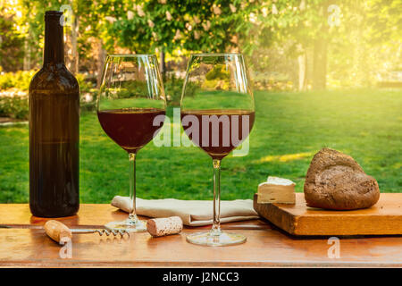 Glasses of red wine and a bottle, with slices of cheese at a picnic, on a wooden table, with green trees and grass in the blurred background, with a r Stock Photo