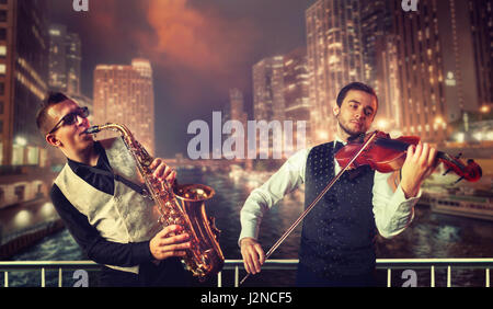 Saxophonist and violinst playing melody against night cityscape background, musical duet. Jazz-man and fiddler Stock Photo