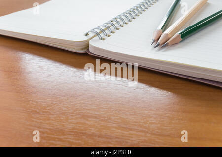 Three pencils on top of open notebook with blank pages on wooden table, selective focus on tips of pencils with blur foreground and background Stock Photo