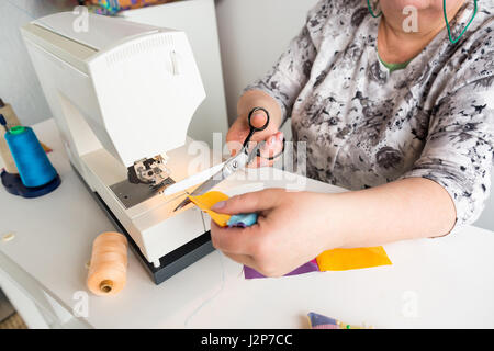 needlework and hand quilting in the workshop of a tailor woman - close up on hands seamstress work on the sewing machine with scissors, she cuts the thread on the stitched colorful fabrics. Stock Photo