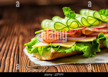 close up of sandwich with ham, cheese, bacon, radish, lettuce, cucumbers and onions on paper, wooden background Stock Photo