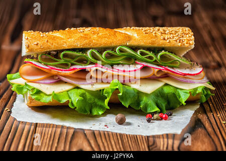 cutting baguette with sesame, ham, cheese, bacon, radish, lettuce, cucumbers and onions decorated dry pepper on dark wooden background Stock Photo