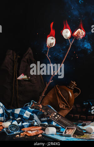 Travelers essentials (maps, knife, flask, backpack and compass) with roasted marshmallows and open flame on a dark background Stock Photo
