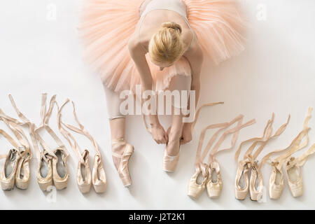 Female ballet dancer sits on the white floor and dresses a beige pointe shoe in the studio. She wears a light dance wear and a peach tutu. On the side Stock Photo