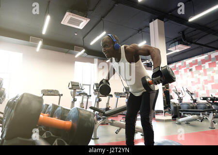 hard workout. Muscular black man wearing sportswear doing exercises with dumbbells for back at gym Stock Photo