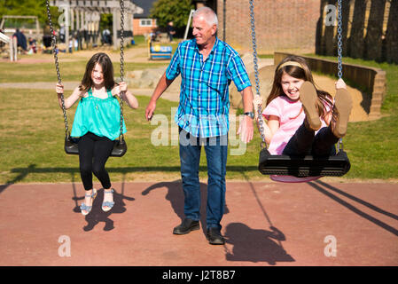 Horizontal portrait of girls being pushed on the swings by their granddad in a park. Stock Photo
