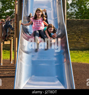 Vertical portrait of two girls sliding down a slide together in the sunshine. Stock Photo