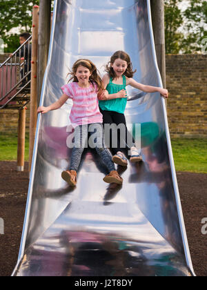 Vertical portrait of two girls sliding down a slide together in the sunshine. Stock Photo