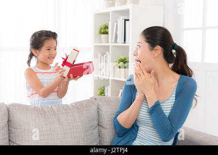 asian chinese mother at the mother's day received a red gift from the little daughter feeling excited and showing emotion sitting on the sofa in the l Stock Photo