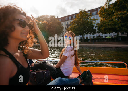Shot of smiling young woman sitting on deck of boat with female friend in front. Two young women on boat in lake. Stock Photo