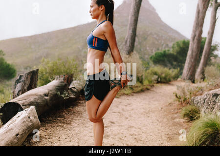 Side view shot of healthy young female runner stretching legs outdoors. Asian woman working out in the morning on mountain trail. Stock Photo