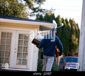 Man on the delivery of parcels with shipped box and a scanner on the belt goes to the house with a glass door surrounded by trees brought to deliver Stock Photo