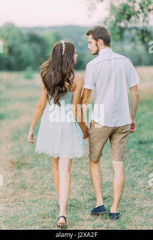 Happy couple holding hands walking through a meadow and lovingly look at each other. Back view. Beautiful summer day Stock Photo