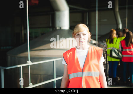 blonde woman in overalls, on the tour of the brewery plant in industrial premises, amid crowds of people in vests, inspecting a factory for training p Stock Photo