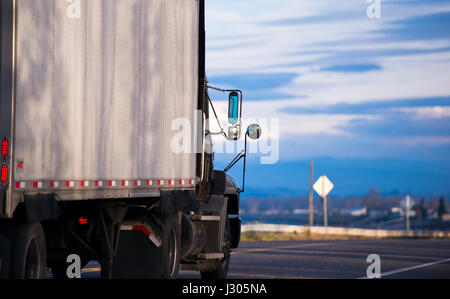 Big black truck with a trailer and a set of rear-view mirrors on the road evening lit by the last rays of the sun against the blue cloudy sky Stock Photo